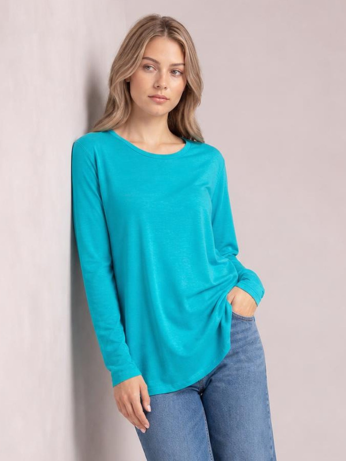 Model wearing a bright turquoise round neck long sleeve top with a relaxed straight silhouette, gently gathered hem, and one hand tucked into a pocket.