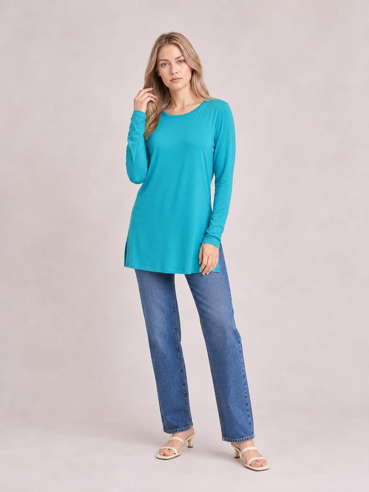 Model poses in a turquoise round neck long sleeve top with a relaxed tunic silhouette, side pockets, and a straight hem over blue jeans.