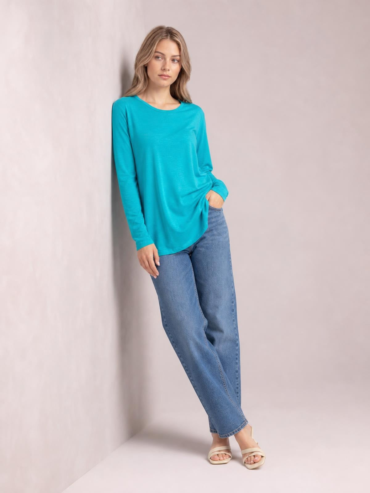 A model wears a turquoise round neck long sleeve top with a relaxed silhouette and curved hem, standing casually with one hand in her jeans pocket.
