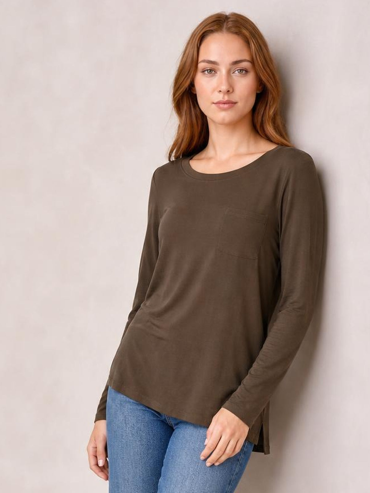 A model wears a brown round neck long sleeve top with a relaxed silhouette, subtle front pocket, and curved hem while standing with one hand at her side.