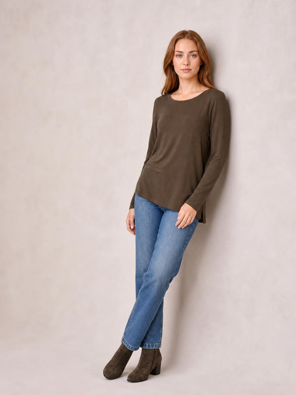 A model poses with crossed legs wearing a brown round neck long sleeve top with a relaxed tunic silhouette, side slits, and a chest pocket.
