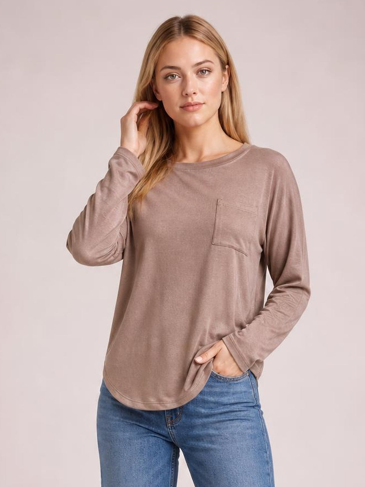 Model poses in a mauve round neck long sleeve top with a relaxed silhouette, curved hem, and visible chest pocket, paired with jeans.