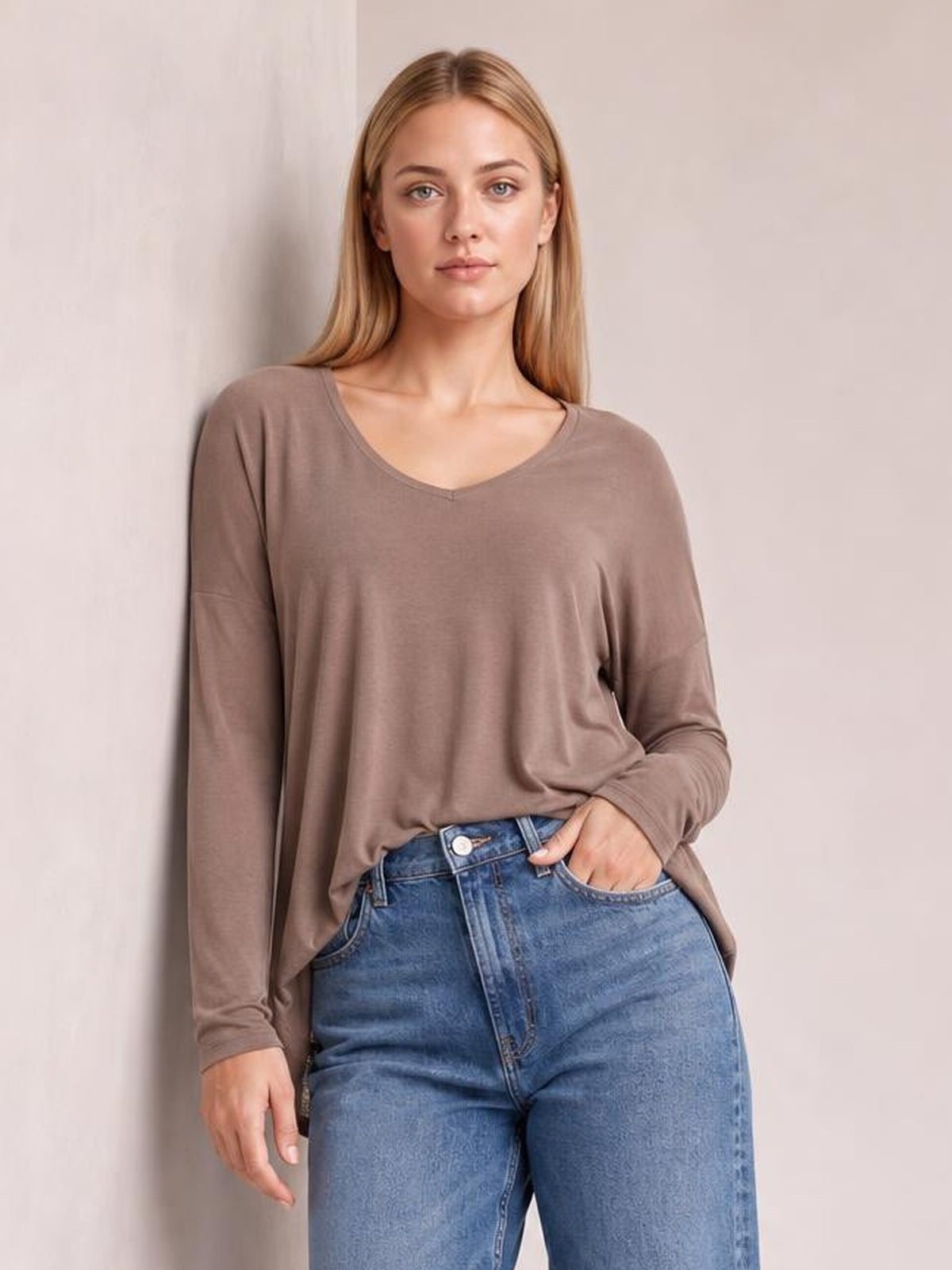 Model wears a taupe long-sleeve top with a softly rounded neckline, relaxed silhouette, and smooth drape, posing with one hand in her jeans pocket.