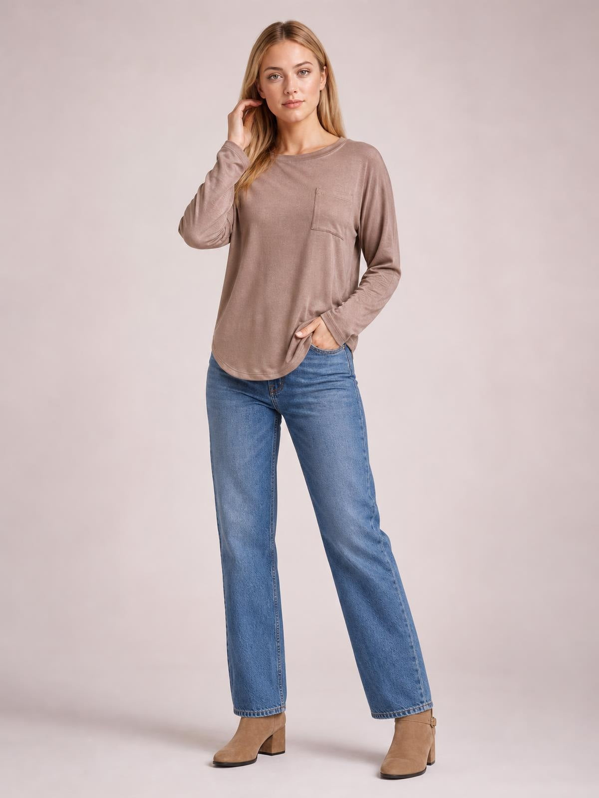 Model wears a taupe round neck long sleeve top with a relaxed silhouette, curved hem, and visible chest pocket, posing with one hand at her hair.
