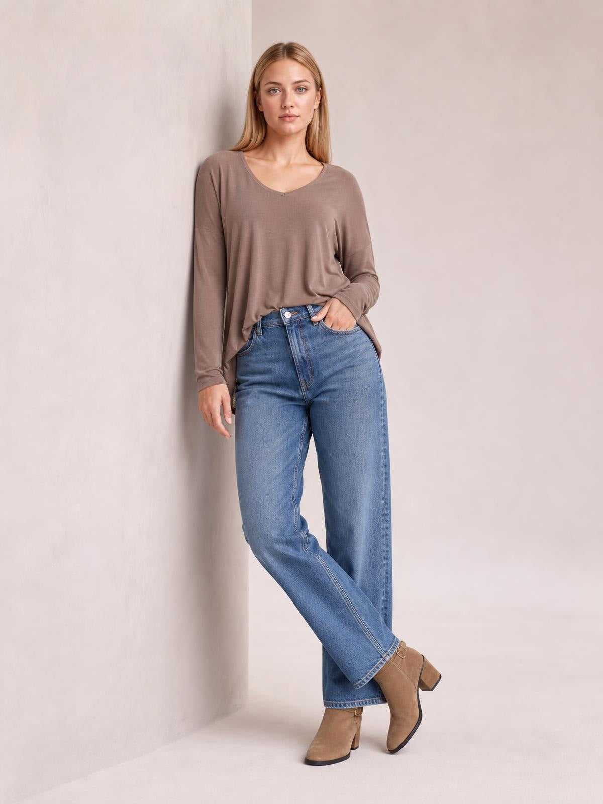 Model wears a taupe long-sleeve top with a soft round neckline and relaxed silhouette, leaning against a wall with crossed legs.