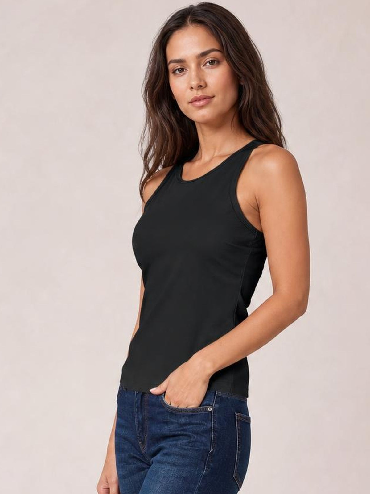 Model wearing a black round neck racerback tank with a fitted silhouette, wide shoulder straps, and a straight hem, posing with one hand in her jeans pocket.