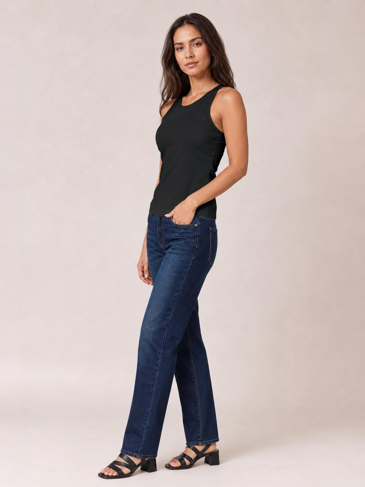 A model wears a black round-neck racerback tank with wide shoulder straps and a smooth, close-fitting silhouette, standing in jeans.