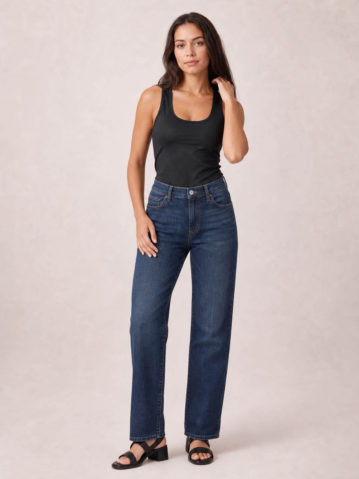 Model wears a black round neck racerback tank with a fitted, slightly stretchy silhouette, paired with jeans, standing with one hand touching her hair.