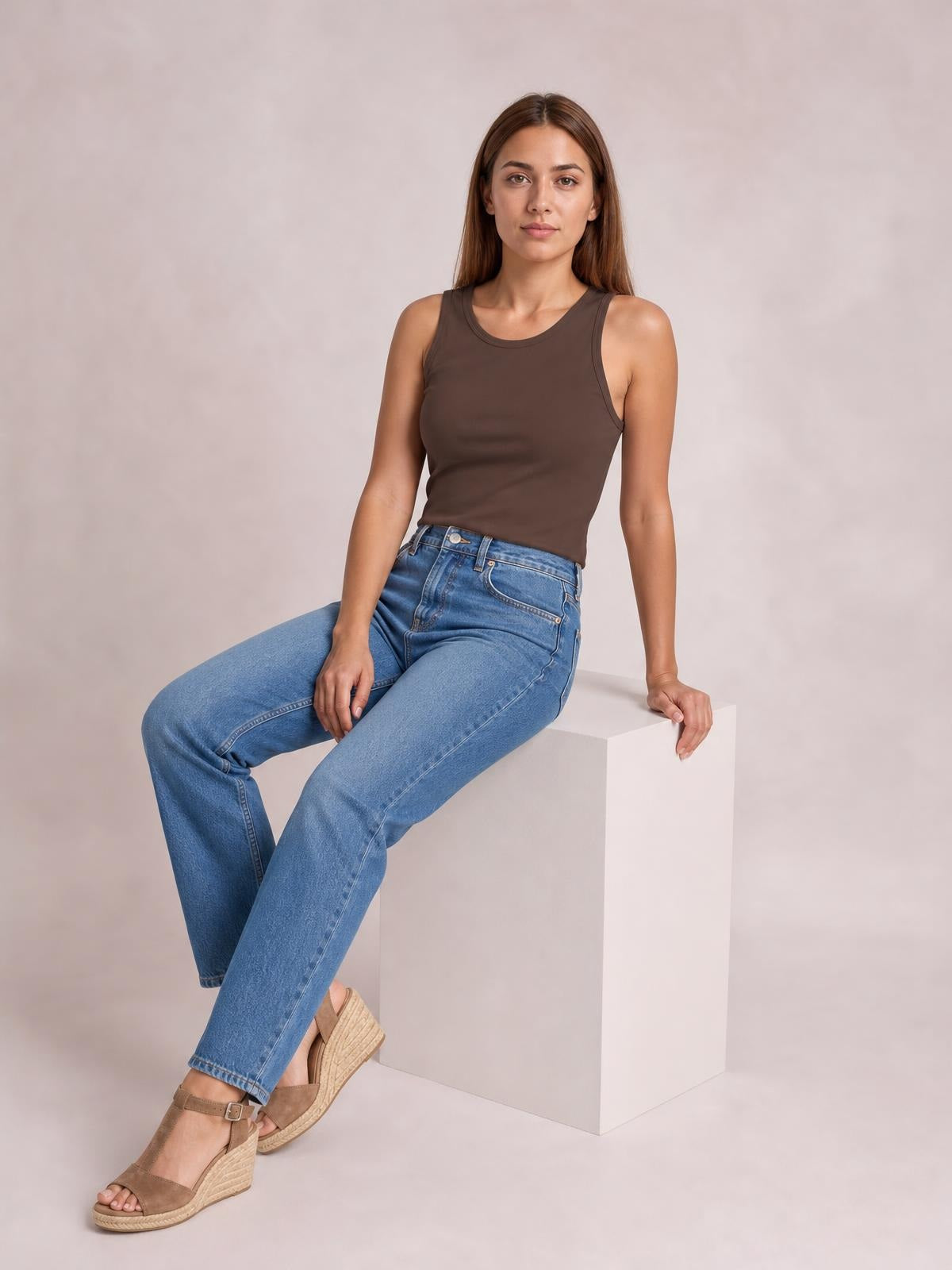 A seated model wears a brown round neck racerback tank with a fitted, sleeveless silhouette and subtly stretchy construction.
