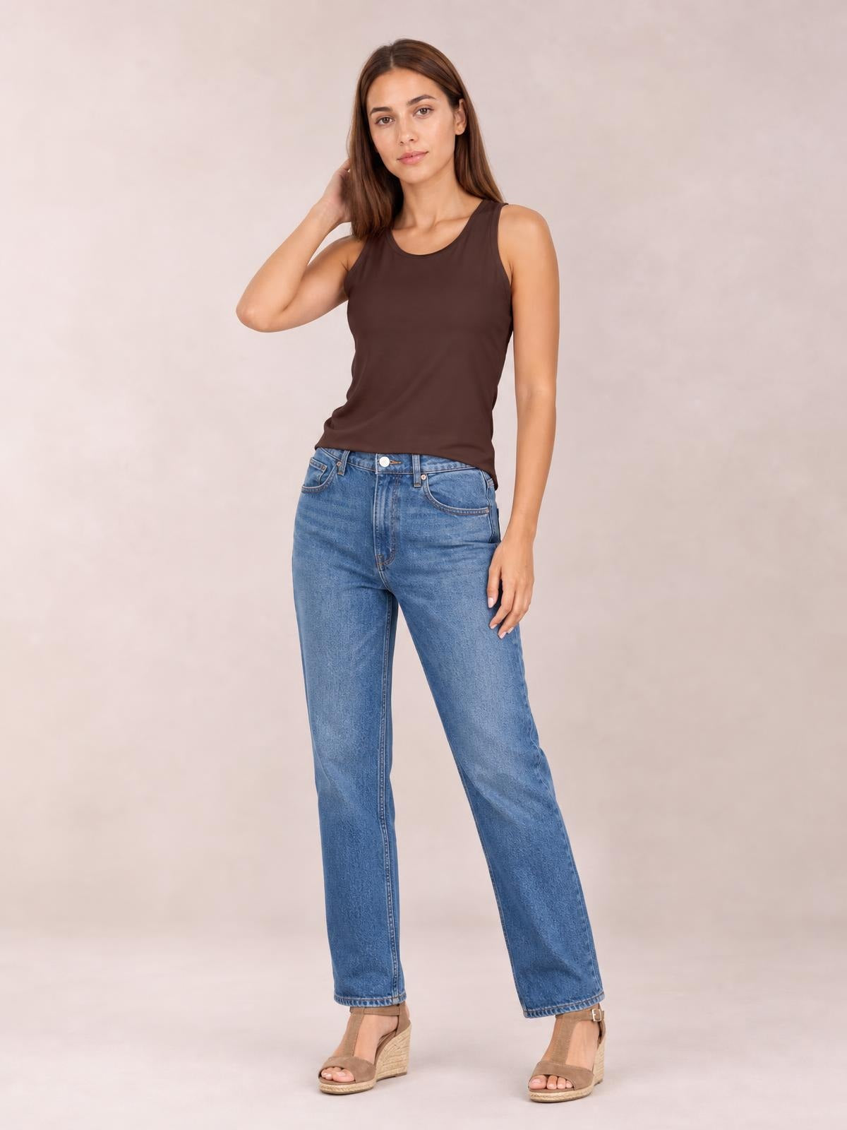 Model poses in a brown round neck racerback tank with wide shoulder straps and a fitted, slightly stretchy silhouette, paired with blue jeans.
