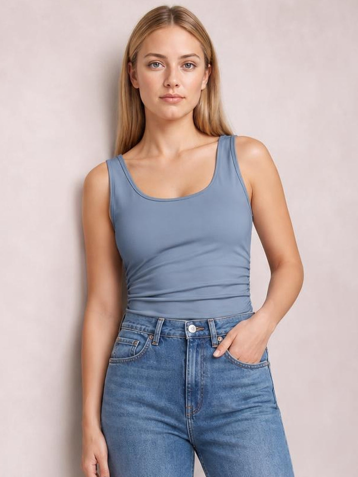 Model wears a slate blue round neck racerback tank with wide straps and a fitted, slightly stretchy silhouette, standing with one hand in her jeans pocket.