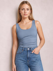 Model wears a slate blue round neck racerback tank with wide straps and a fitted, slightly stretchy silhouette, standing with one hand in her jeans pocket.