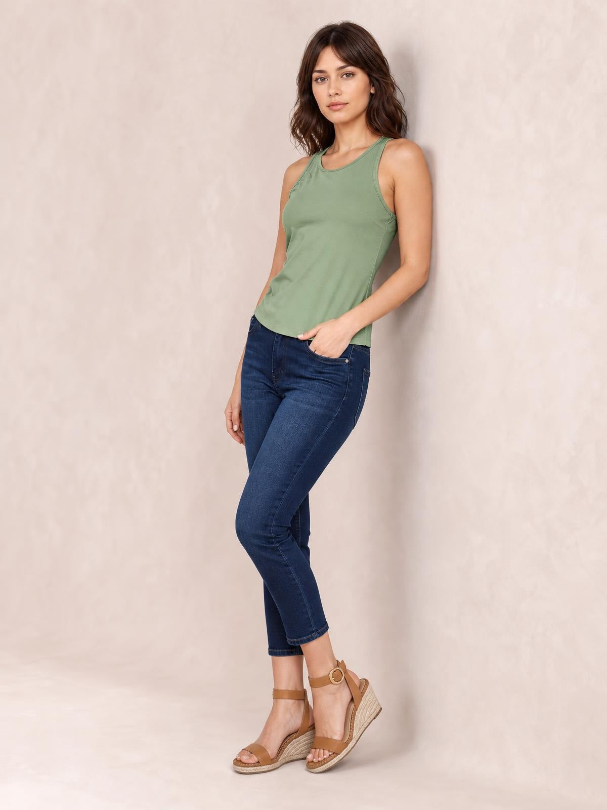 Model wearing a sage green round neck racerback tank with wide shoulder straps and a slim, slightly stretchy silhouette, posing with one hand in her jeans pocket.
