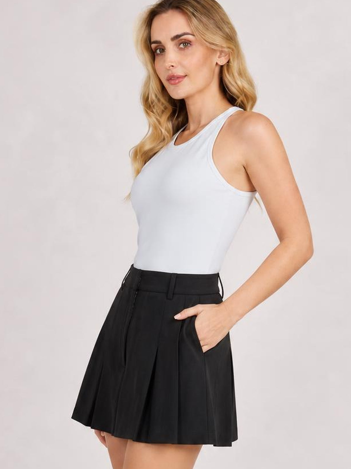 Model wearing a pale blue round neck racerback tank with wide shoulder straps, sleeveless cut, and a smooth, fitted silhouette tucked into a black pleated skirt.
