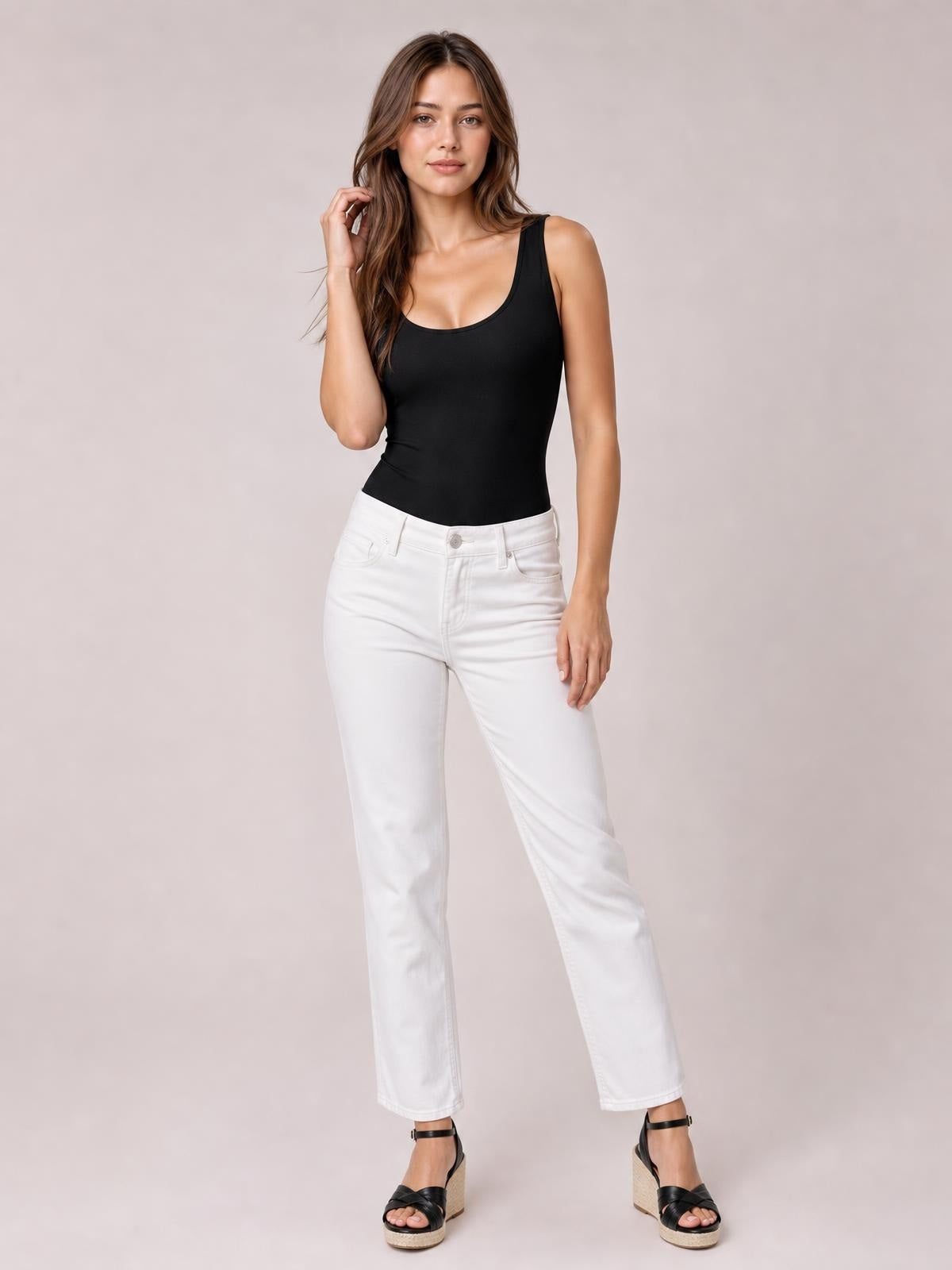 Model poses standing in a black round-neck bodysuit with wide shoulder straps and a smooth, fitted silhouette tucked into white jeans.