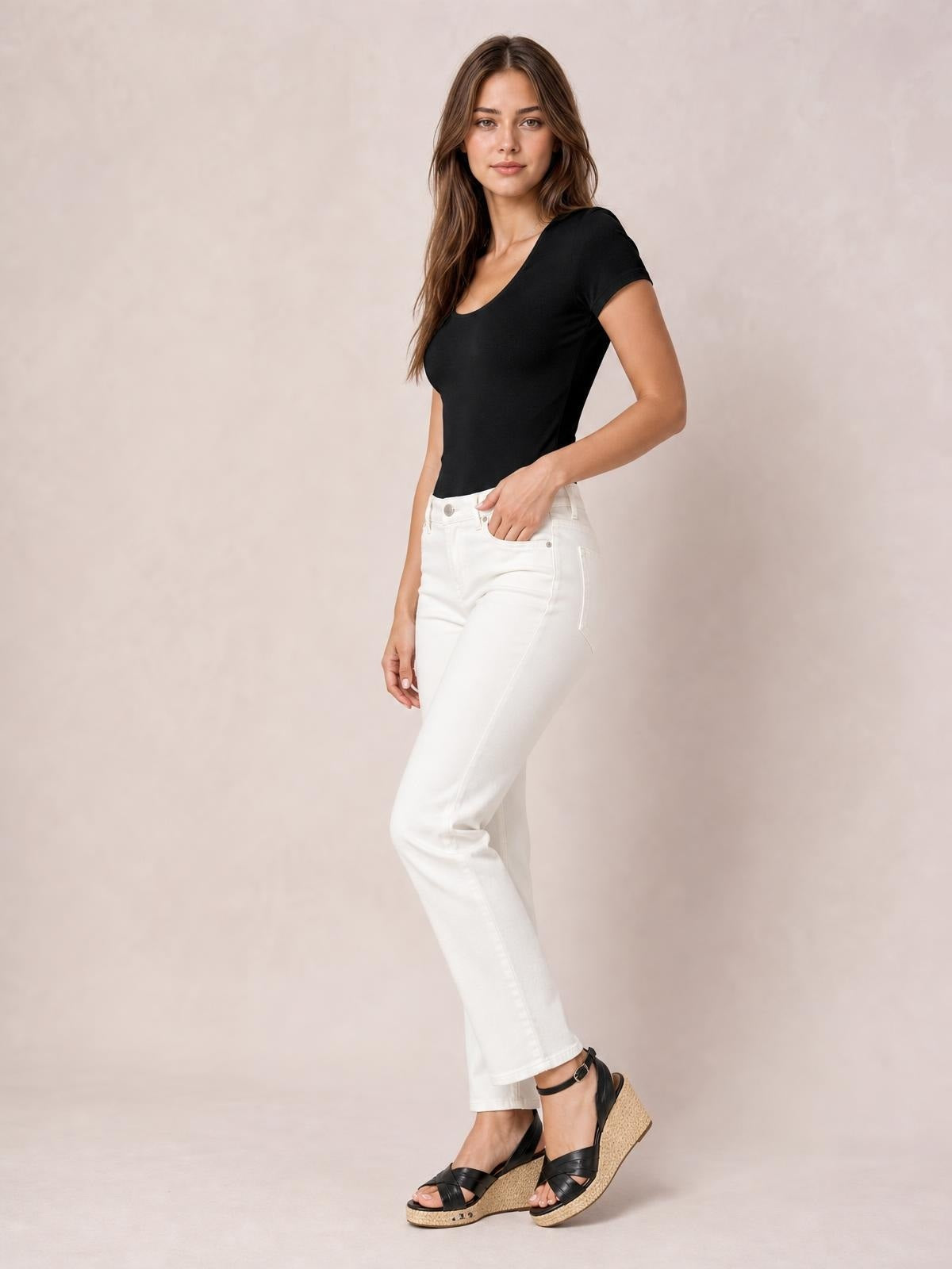 Model wears a black round neck short sleeve bodysuit with a fitted silhouette and smooth basic construction, posing with one hand in her jeans pocket.