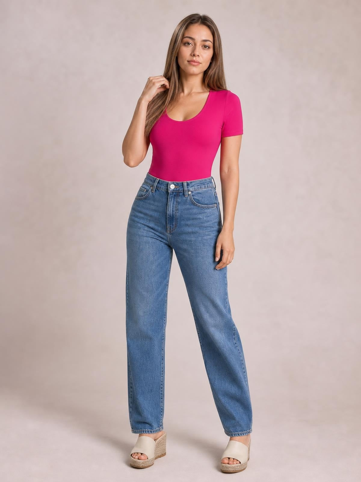 Model poses in a bright pink round neck short sleeve bodysuit with a fitted silhouette and smooth construction, paired with high-waisted blue jeans.