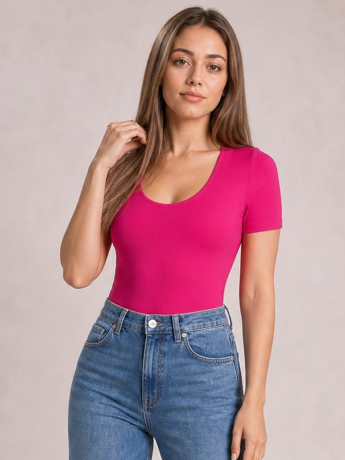 A model wears a bright pink round-neck short-sleeve bodysuit with a close-fitting silhouette, lightly touching her hair in a relaxed pose.