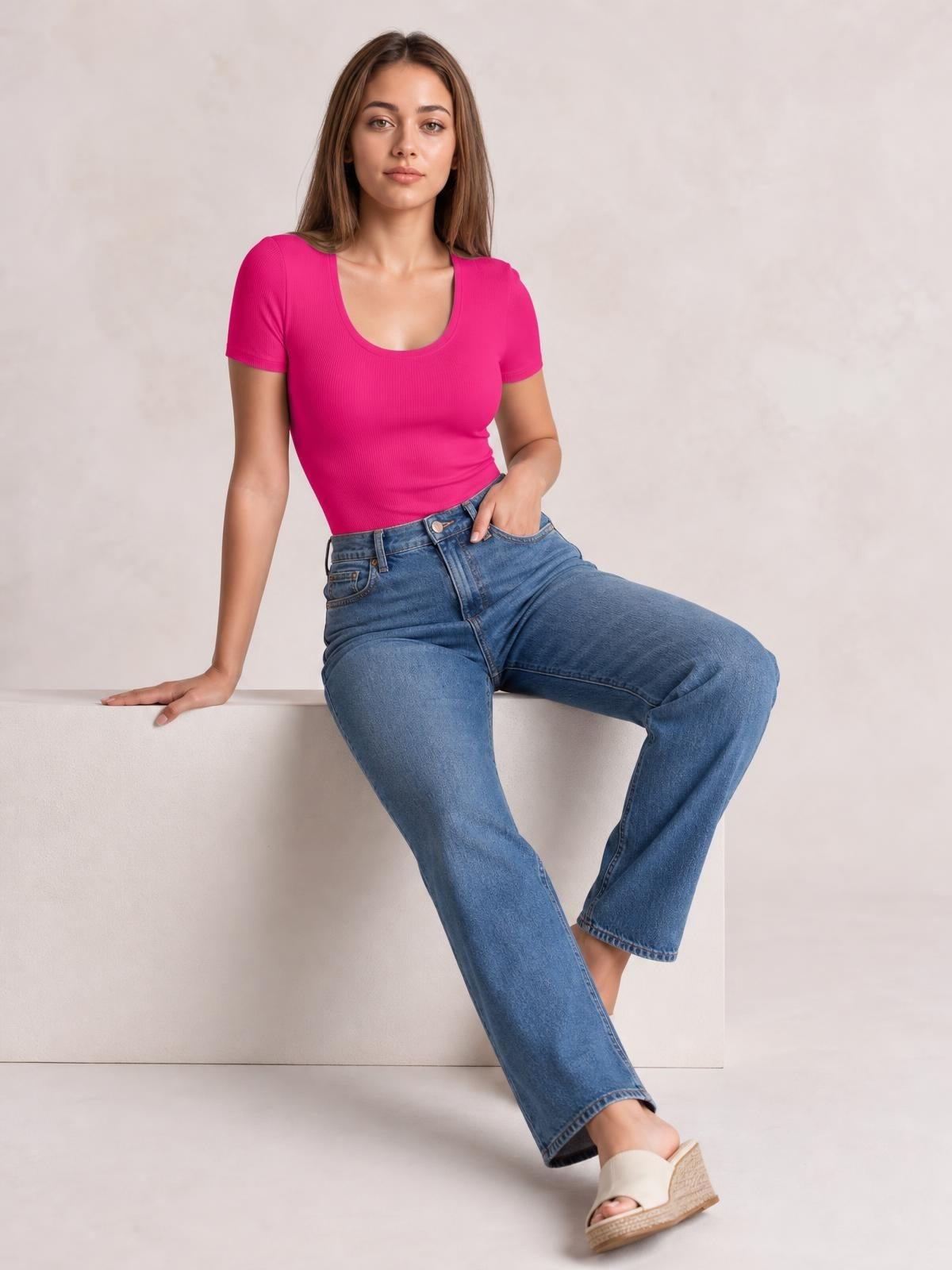 Model seated in a bright pink round neck short sleeve bodysuit with a fitted silhouette and ribbed construction, paired with blue jeans.