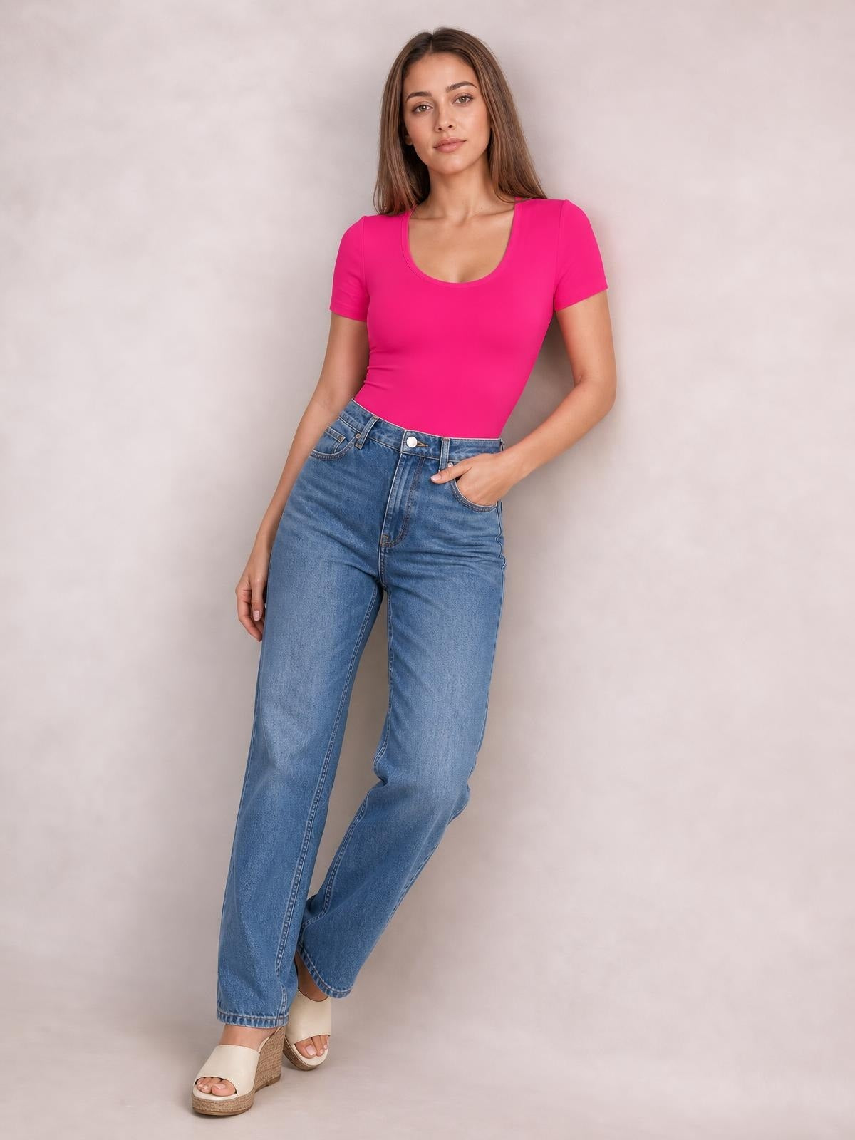 Model poses in a bright pink round neck short sleeve bodysuit with a fitted silhouette, smooth construction, and moderate stretch, paired with jeans.