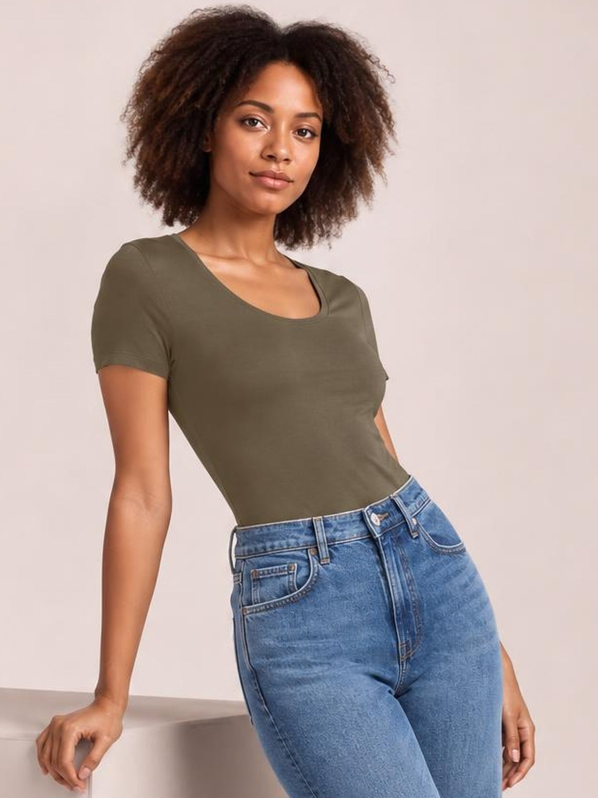 Model wearing an olive round neck short sleeve bodysuit with a fitted, smooth silhouette and moderate stretch, leaning casually with one hand supported.