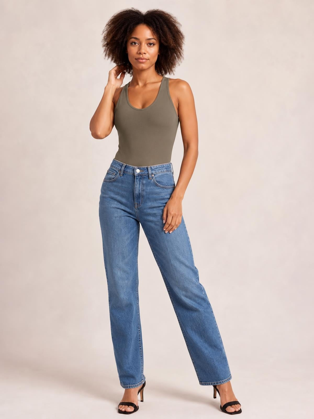 Model wears an olive round-neck bodysuit with a fitted silhouette and wide shoulder straps, standing in jeans with one hand touching her hair.