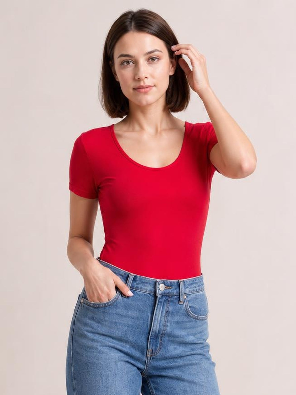 Model wears a red round-neck short-sleeve bodysuit with a fitted silhouette, smooth opaque construction, and one hand resting in her jeans pocket.