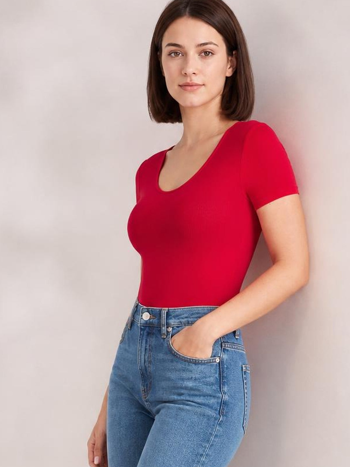 Model wears a red round neck short sleeve bodysuit with a smooth, fitted silhouette, moderate stretch, and clean construction, posing with one hand in her jeans pocket.