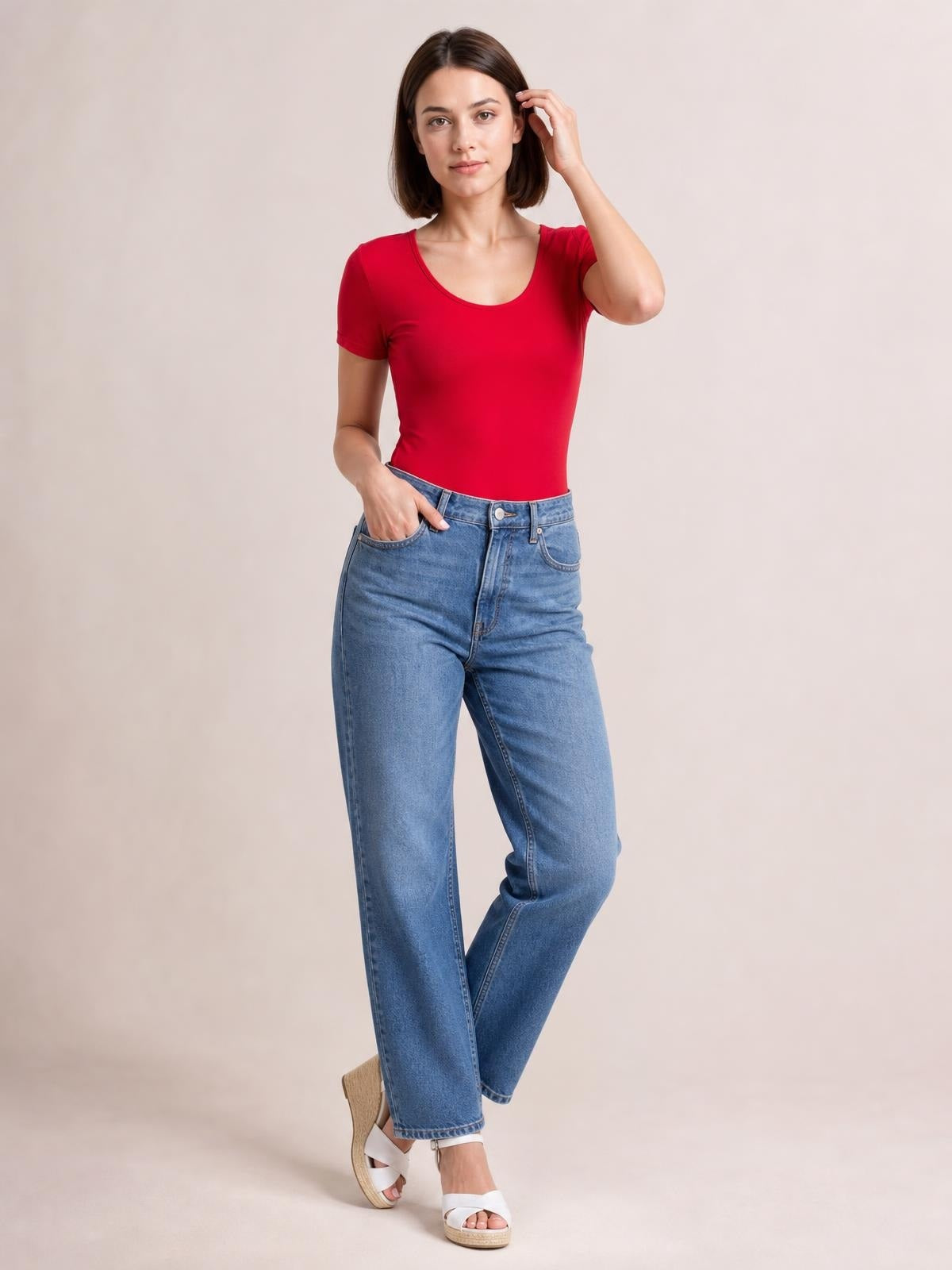 Model wearing a fitted red round neck short sleeve bodysuit with a smooth basic silhouette, posing with one hand in her jeans pocket and the other touching her hair.