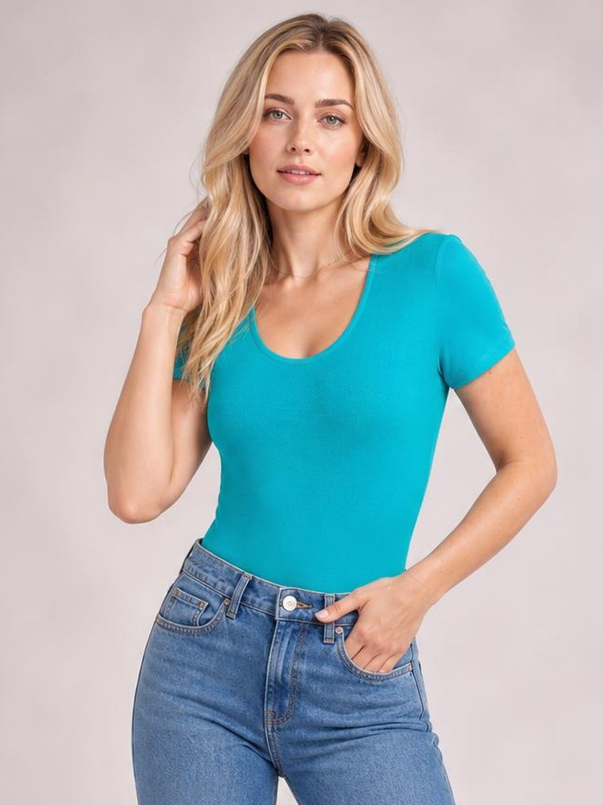 Model wears a fitted turquoise round-neck short-sleeve bodysuit with a smooth opaque finish, styled with blue jeans and one hand resting in a pocket.