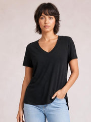 Model wearing a black V-neck high-low T-shirt with short sleeves, relaxed silhouette, and visible side slit, posing with one hand in her jeans pocket.