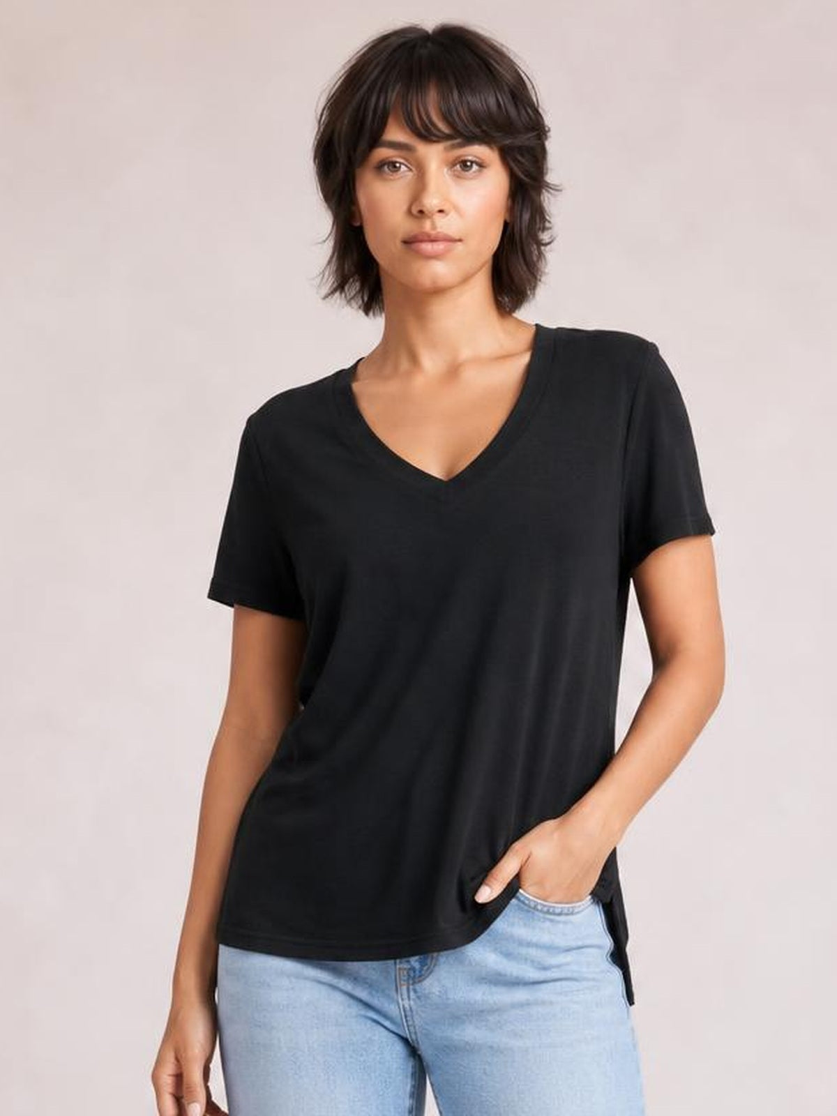 Model wearing a black V-neck high-low T-shirt with short sleeves, relaxed silhouette, and visible side slit, posing with one hand in her jeans pocket.