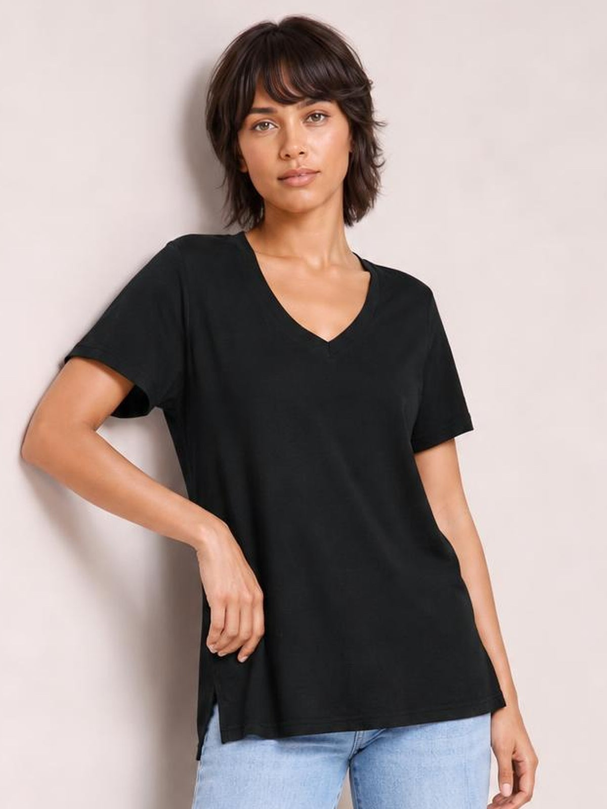 Model poses in a black V-neck high-low T-shirt with short sleeves, relaxed silhouette, and visible side slit, paired with light-wash jeans.