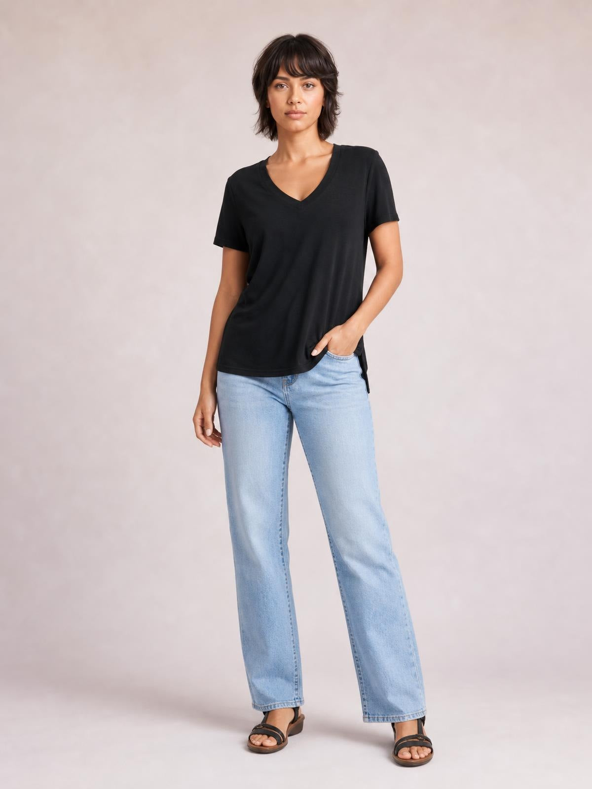 Model wears a black V-neck high-low T-shirt with short sleeves and a relaxed silhouette, standing with one hand in her jeans pocket.