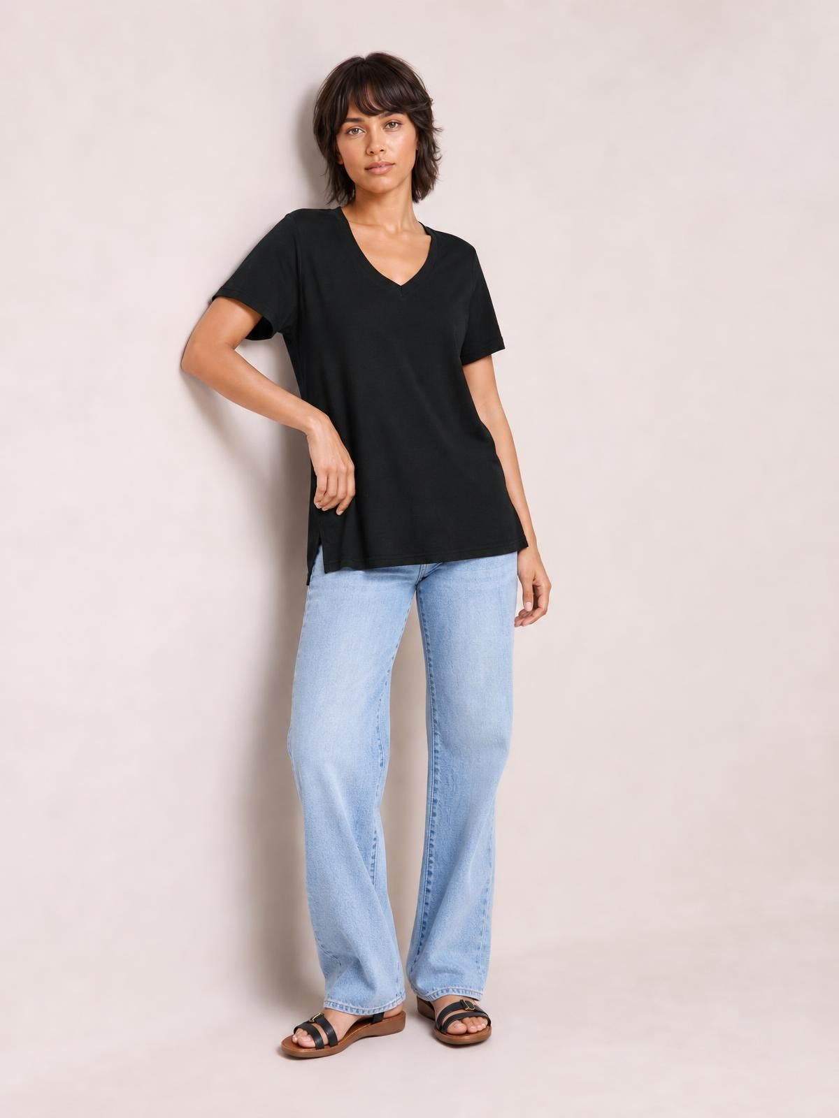 Model poses in a black V-neck high-low T-shirt with short sleeves, relaxed silhouette, and visible side slit, paired with light-wash jeans.