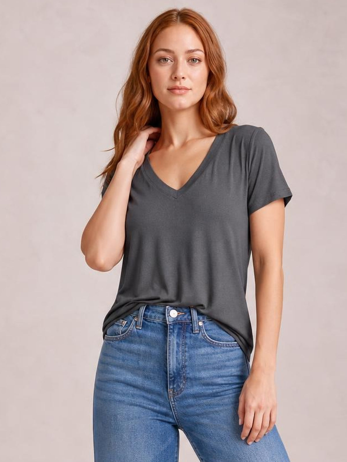 Model wears a charcoal gray V-neck high-low T-shirt with short sleeves, a relaxed draped silhouette, and side slit, posing with one hand near her hair.