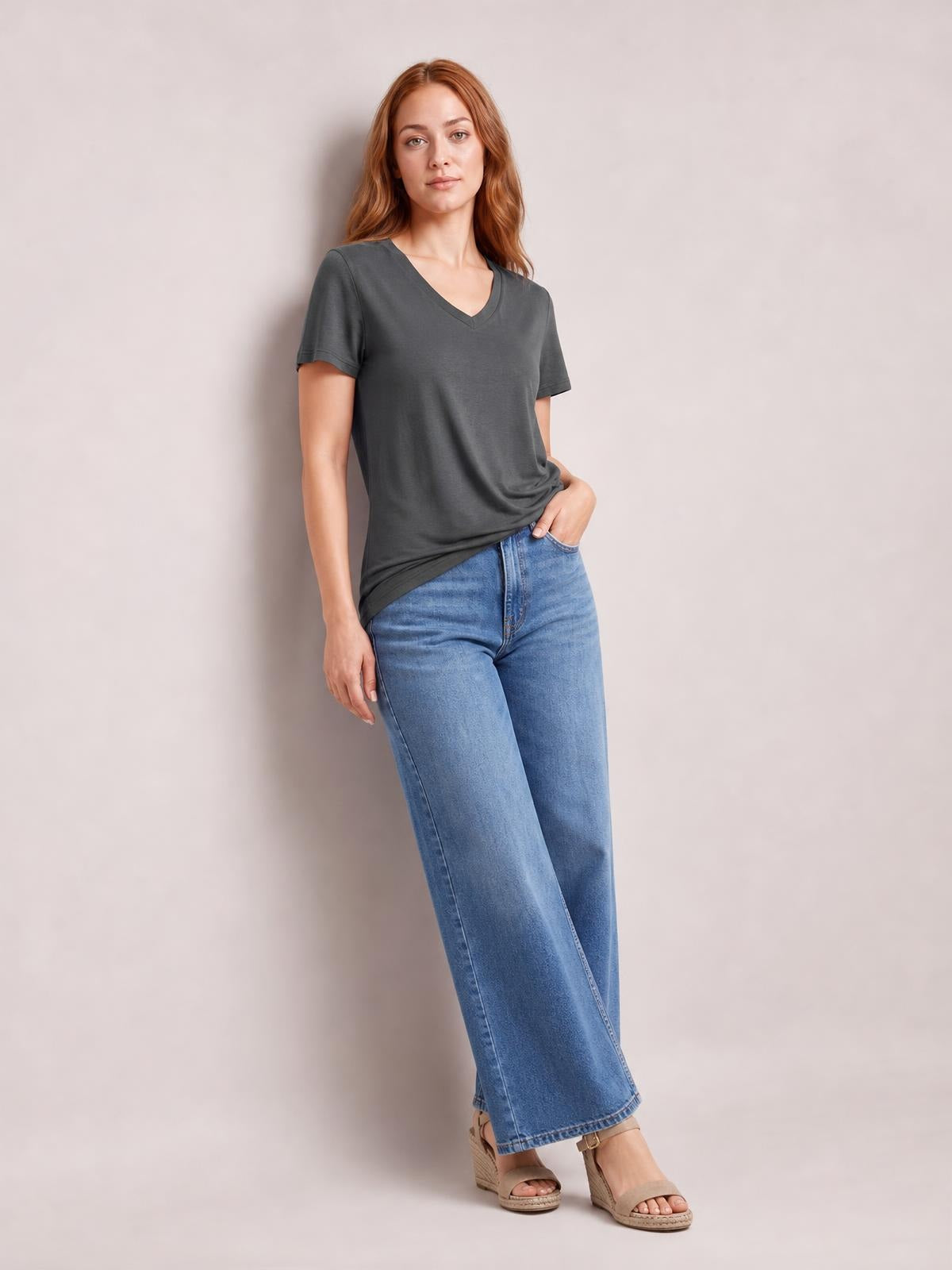 Model poses in a charcoal V-neck T-shirt with short sleeves, relaxed silhouette, curved high-low hem, and side slit, styled with jeans.