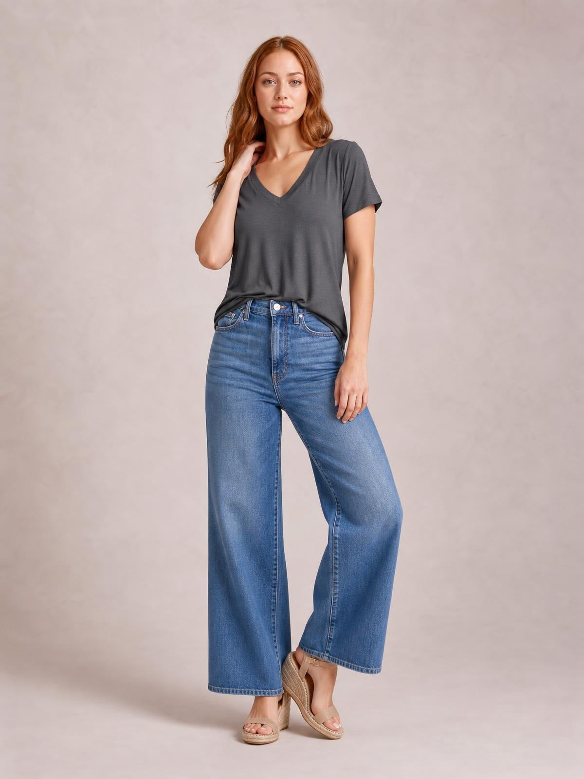 Model poses in a charcoal V-neck high-low T-shirt with short sleeves, relaxed silhouette, and side slit, styled with wide-leg jeans.