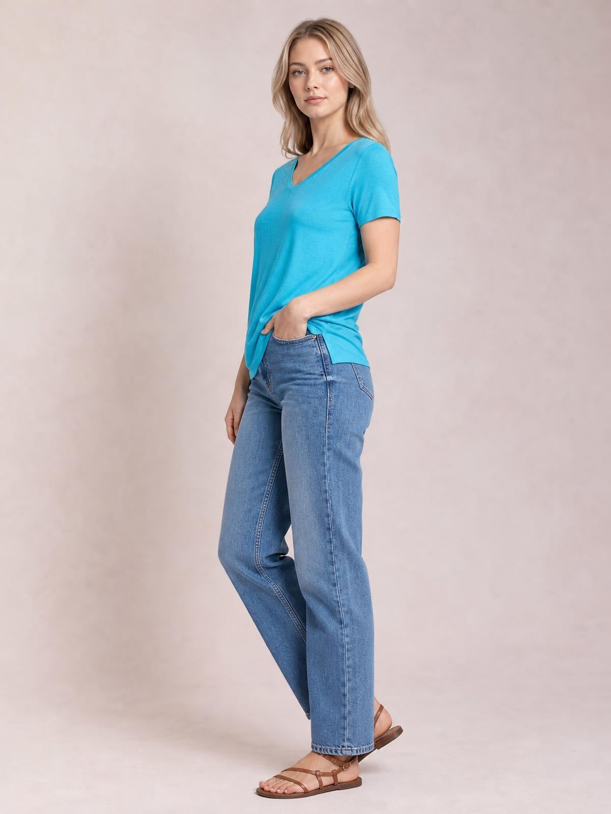Model wears a turquoise V-neck high-low T-shirt with short sleeves and a side slit, styled in a relaxed silhouette while posing with one hand in her jeans pocket.