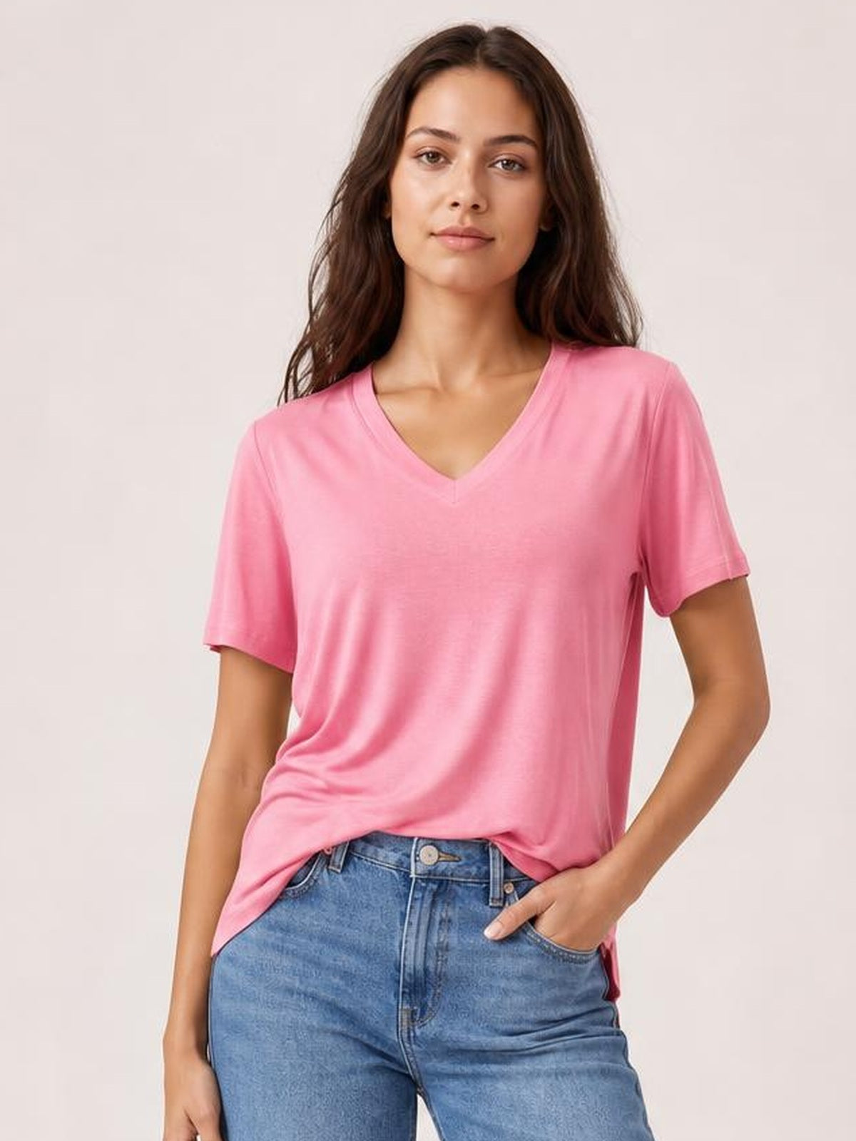 Model wearing a pink V-neck high-low T-shirt with short sleeves, relaxed silhouette, soft drape, and visible side slit, paired with jeans.