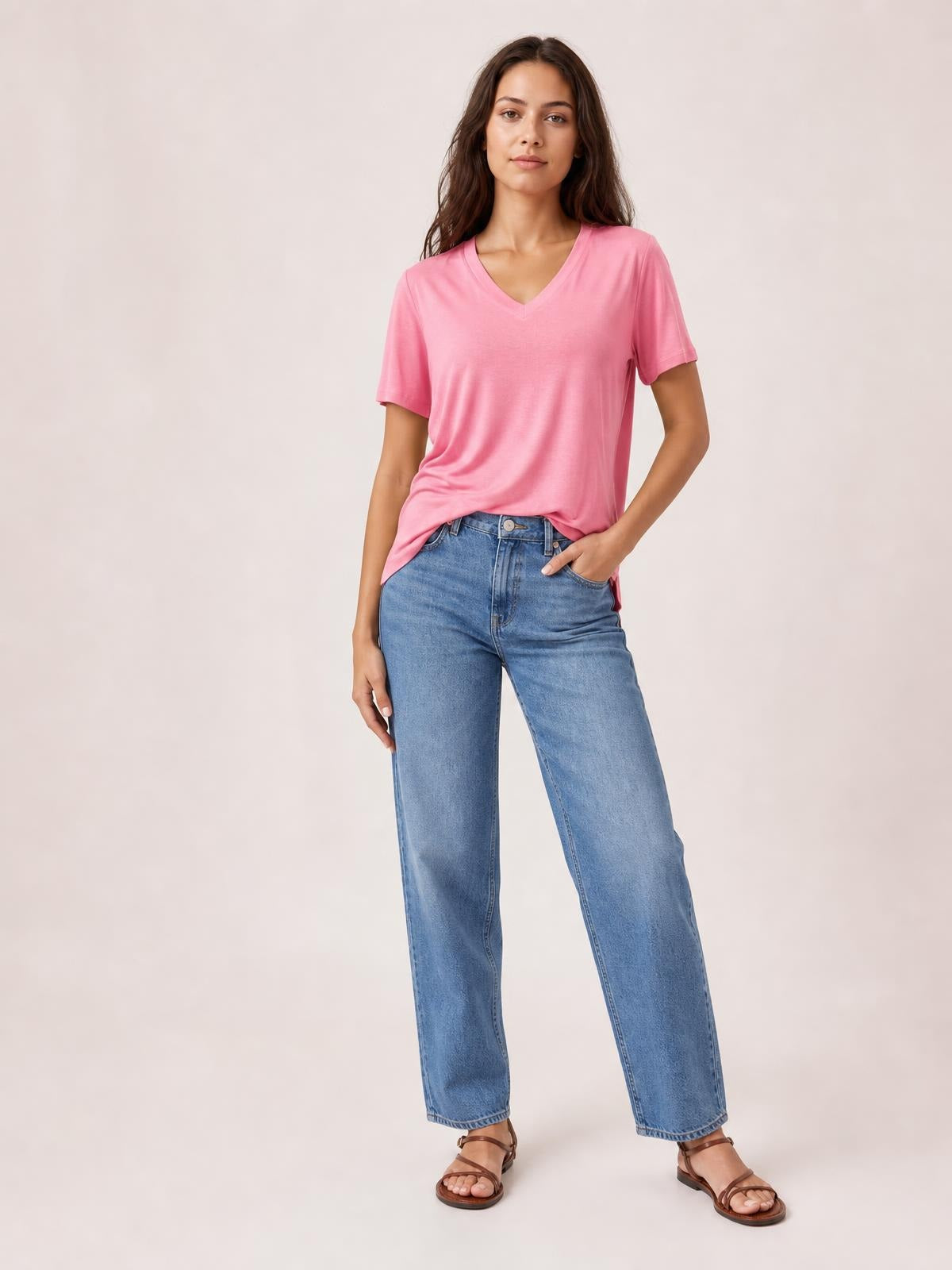 Model wearing a pink V-neck high-low T-shirt with short sleeves, relaxed silhouette, and side slit, posed with one hand in her jeans pocket.