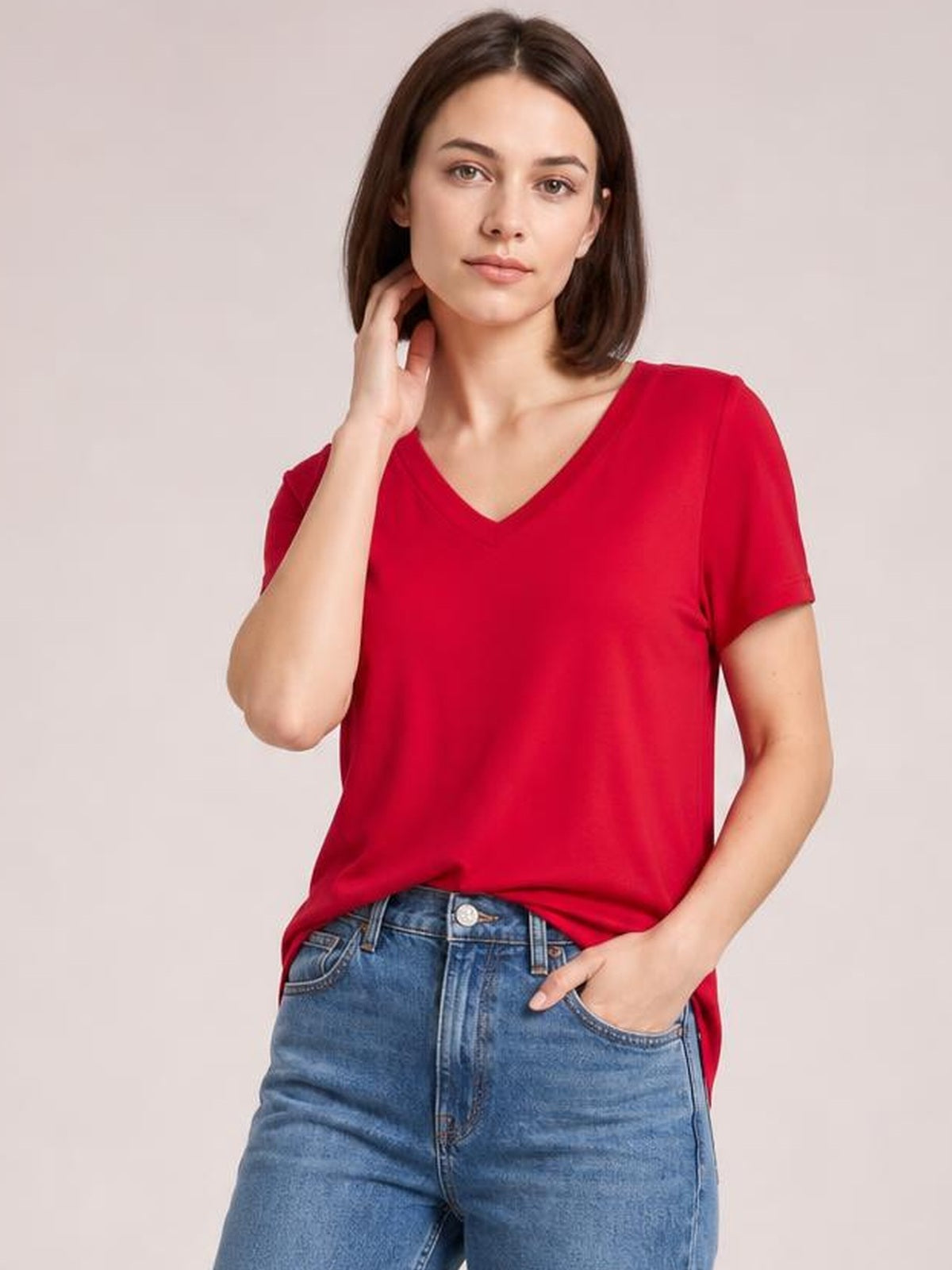 Model wearing a bright red V-neck high-low T-shirt with short sleeves, relaxed silhouette, and side slit, posing with one hand in her jeans pocket.