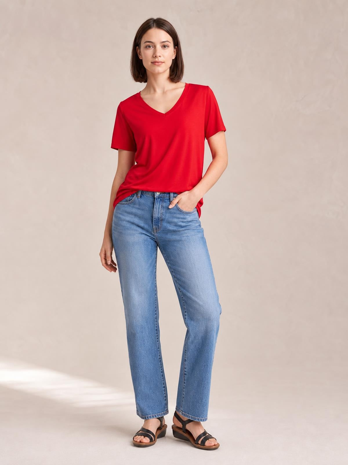 Model wearing a red V-neck high-low T-shirt with short sleeves, relaxed drape, and side slit, standing with one hand in her jeans pocket.