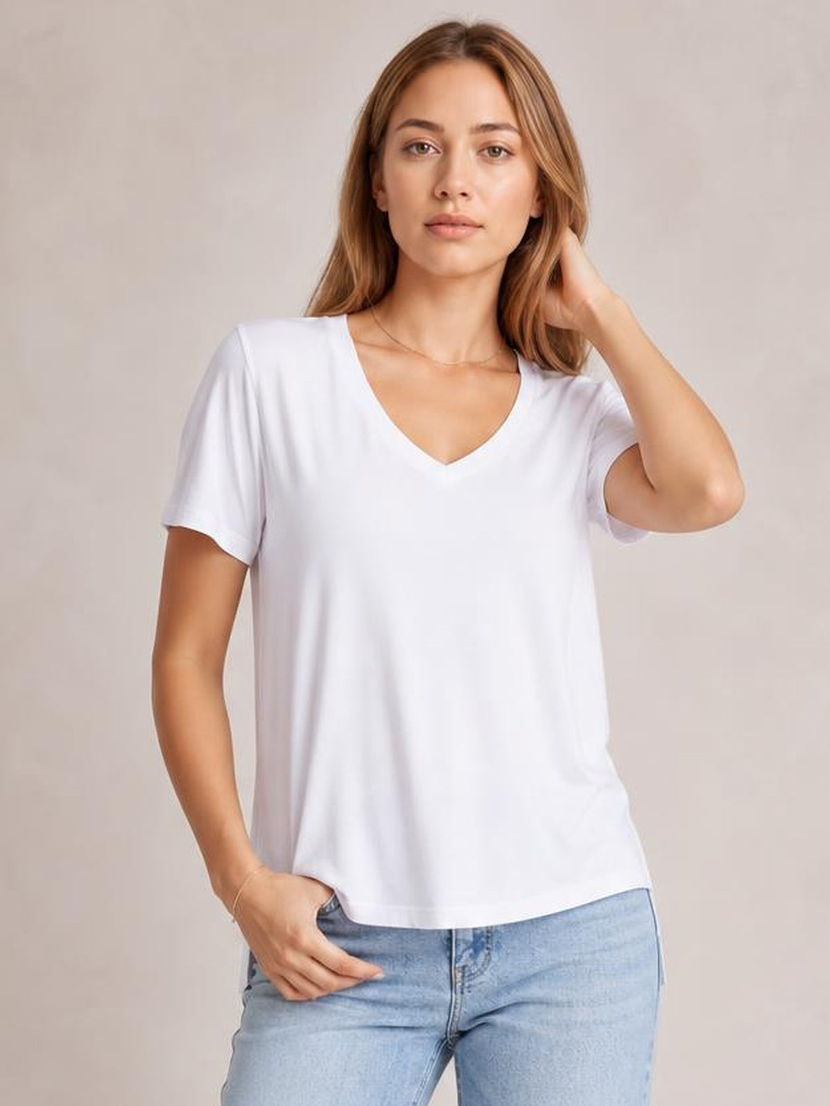 A model poses in a white V-neck high-low T-shirt with short sleeves, a relaxed silhouette, and a curved hem while resting one hand near her hair.