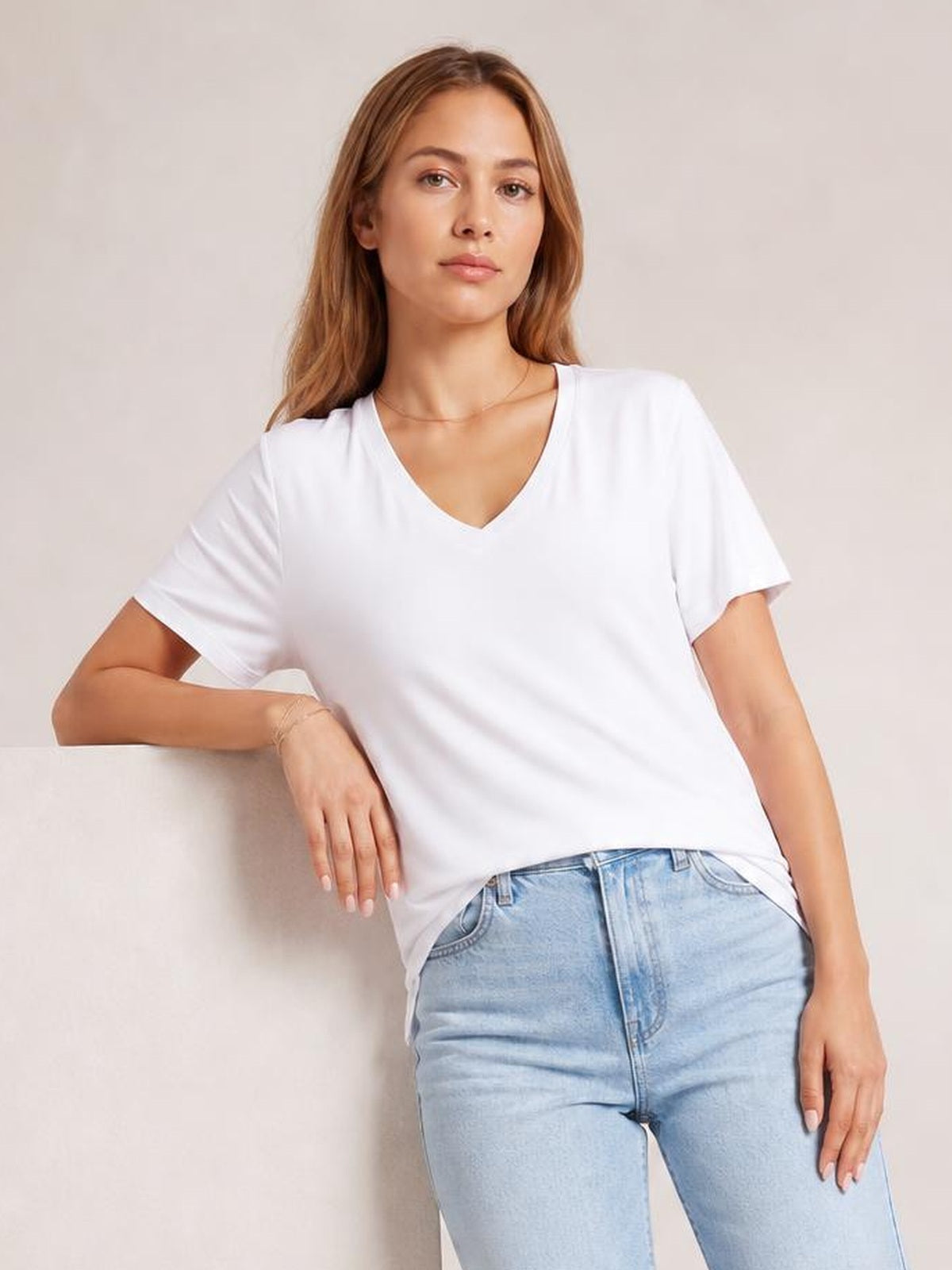 A model leans casually in a white V-neck high-low T-shirt with short sleeves, relaxed silhouette, and a visible side slit at the hem.