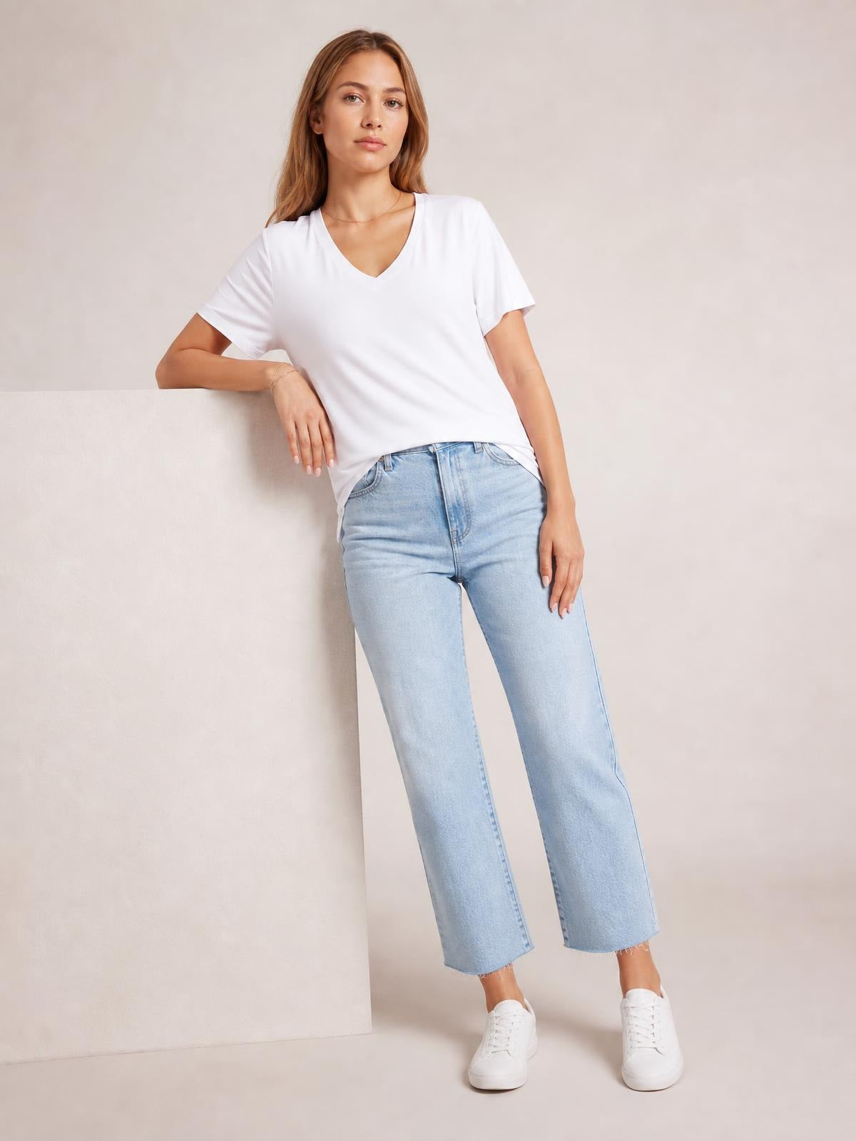 Model leaning with one arm resting, wearing a white V-neck high-low T-shirt with short sleeves, relaxed silhouette, and visible side slit.