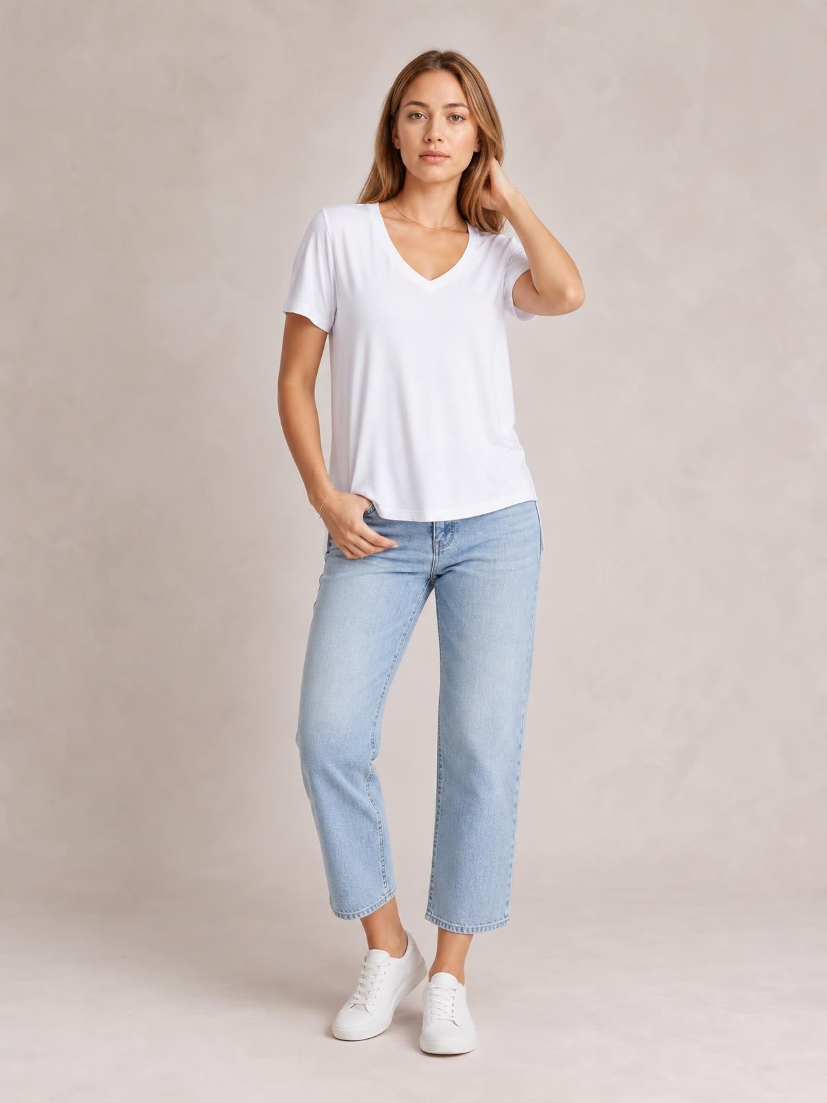 A model poses in a white V-neck high-low T-shirt with short sleeves, relaxed silhouette, curved hem, and subtle side slit, paired with jeans.