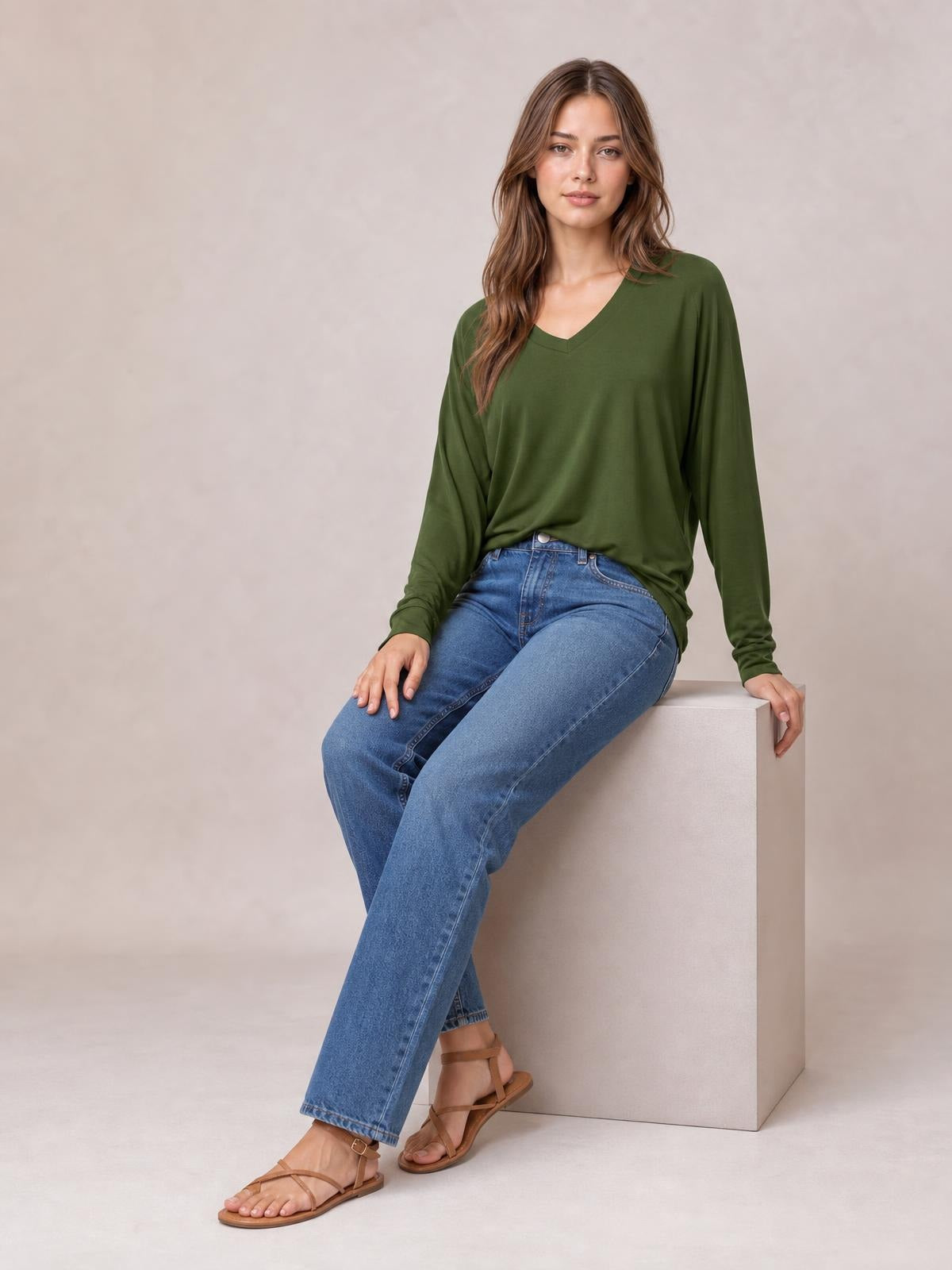 Model seated in a relaxed olive-green V-neck top with long lantern sleeves, gathered cuffs, and a loose, draped silhouette styled with blue jeans.