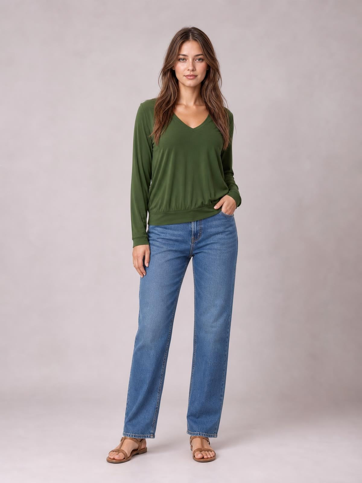 Model wears an olive green V-neck top with long lantern sleeves, a relaxed silhouette, and a banded hem, posing with one hand in her jeans pocket.