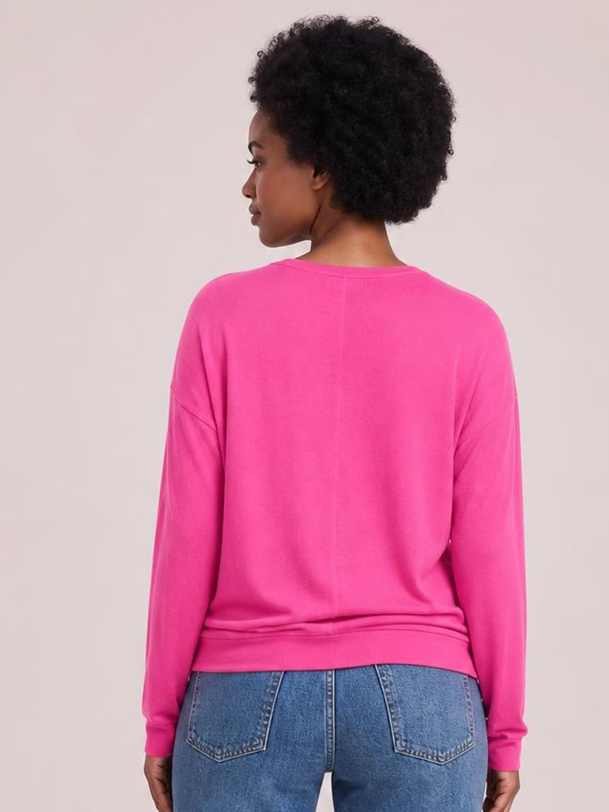 A model stands facing away in a bright pink long-sleeve top with a relaxed silhouette, dropped shoulders, ribbed cuffs, hem, and center-back seam.